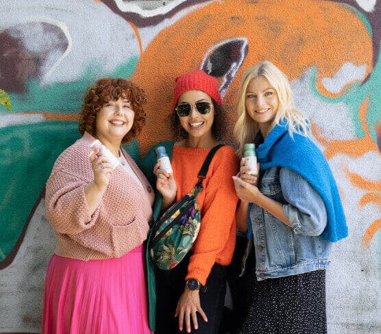 Na zdjęciu znajduję się jeden z kadrów sesji zdjęciowej wykonanej dla marki Collibre. Na tle kolorowego graffiti stoją trzy uśmiechnięte kobiety. Modelki w dłoniach trzymają butelki z kolagenem.