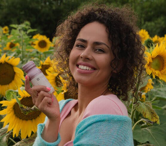 Na zdjęciu znajduję się jeden z kadrów sesji zdjęciowej wykonanej dla marki Collibre. Na tle kolorowego pola słonecznikowego stoi uśmiechnięta modelka. Kobieta w dłoni trzyma butelkę kolagenu.
