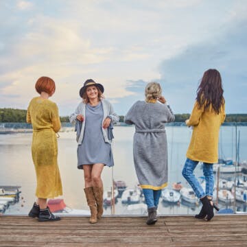 Zdjęcie przedstawia jednego z klientów Agencji Detalle - markę You by Tokarska. Na fotografii znajdują się 4 modelki stojące na tarasie. W tle widać taflę jeziora. Trzy kobiety stoją tyłem, jedna z nich - właścicielka jest odwrócona przodem i patrzy przed siebie. Wszystkie kobiety mają na sobie swetry marki You by Tokarska. 