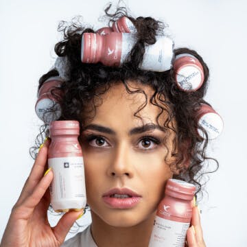 Zdjęcie przedstawia jednego z klientów Agencji Detalle - markę Collibre. Na portretowej fotografii znajduję się kobieta w kręconych włosach. W kosmykach jej włosów i w dłoniach znajdują się małe butelki z kolagenem.